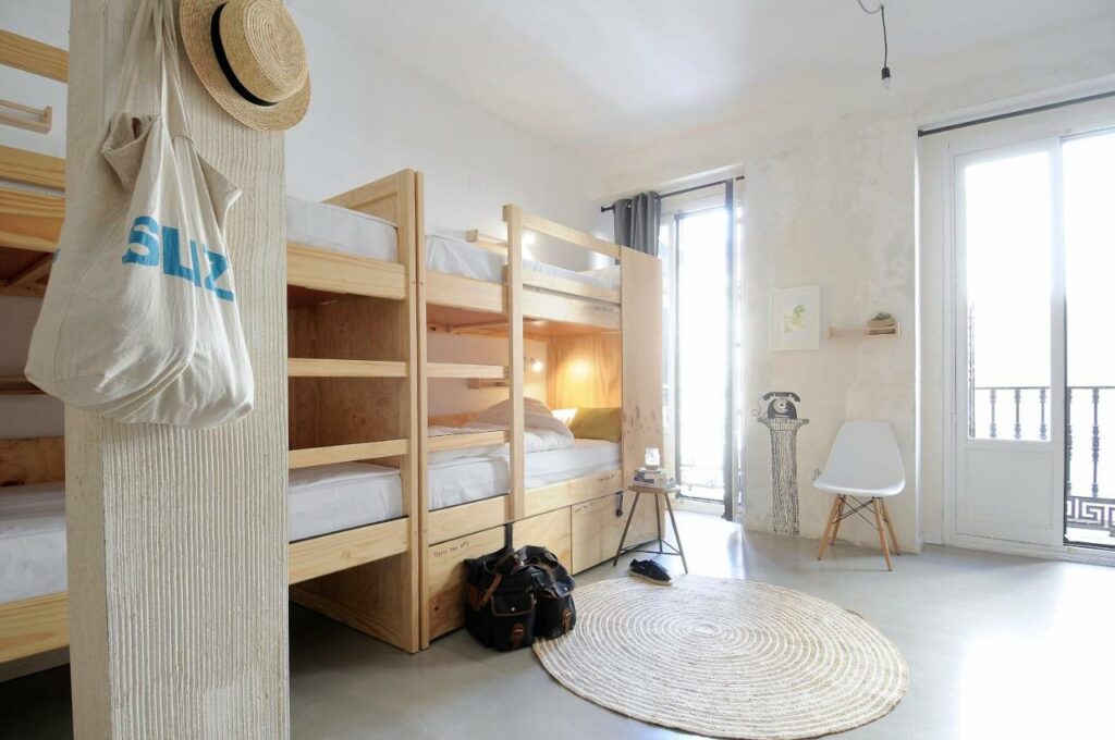 Quarto do The Hat Madrid, uma das recomendações de hostels em Madri. Beliches de madeira clara estão no lado esquerdo do quarto com bolsas e um chapéu pendurados. Há um tapete no centro do quarto, e a parede do fundo tem duas janelas grandes com uma cadeira entre elas.