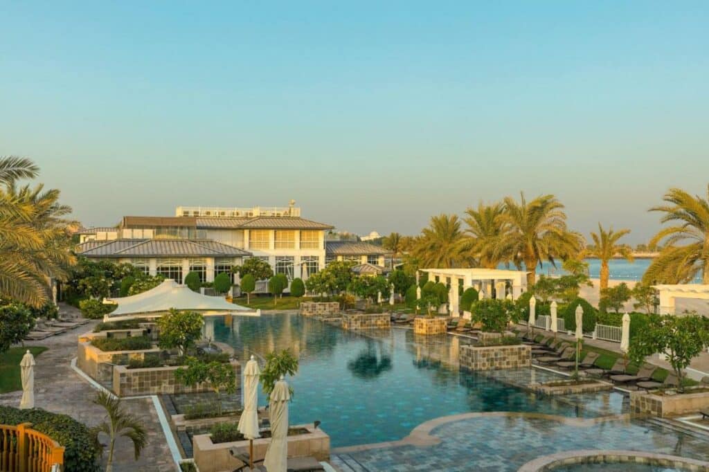 Piscina ao ar livre do The St. Regis Abu Dhabi. Uma piscina no meio, ao redor árvores e cadeiras de praia. No fundo o hotel e no lado direito palmeiras e a praia.