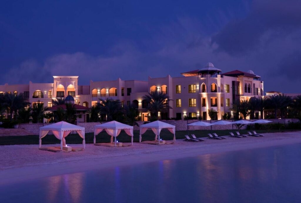 Área externa do Traders Hotel, Abu Dhabi. O mar no lado direito, do lado esquerdo tendas e guarda-sóis com cadeira de praia, palmeiras no fundo e atrás o hotel iluminado com luz amarela.
