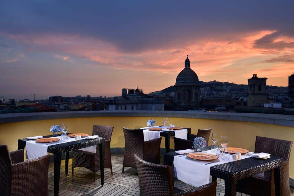 Parte do UNAHOTELS Napoli ao ar livre com 3 mesas e cada mesa com duas cadeiras e a vista do terraço para as construções da cidade, imagem ilustrando post Hotéis em Nápoles.