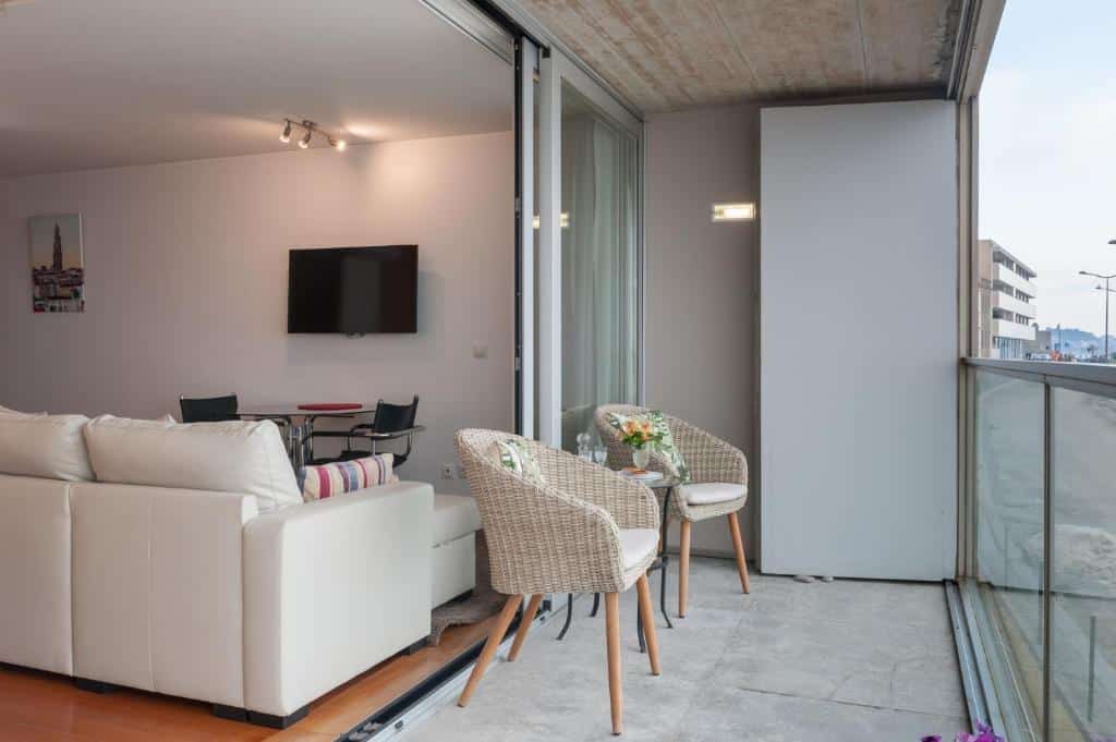 Varanda do Bartolomeu Beach Apartments com duas poltronas na sacada e ao fundo uma sala de estar com sofá e TV.