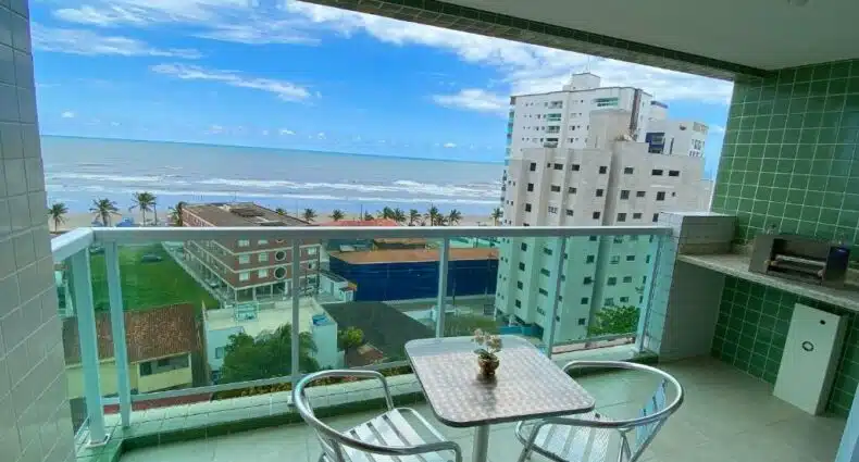 Varanda de apartamento no Edifício Baalbek com mesa e duas cadeiras de metal centralizadas, pequena churrasqueira no canto direito e vista para a praia, para representar pousadas em Mongaguá.