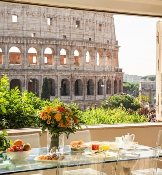 vista da sala do 47Luxury Suites - Colosseo, um aluguel de temporada em Roma, com café posto à mesa de vidro, cadeiras e vaso de flores, dando vista para o Coliseu e plantinhas ao redor