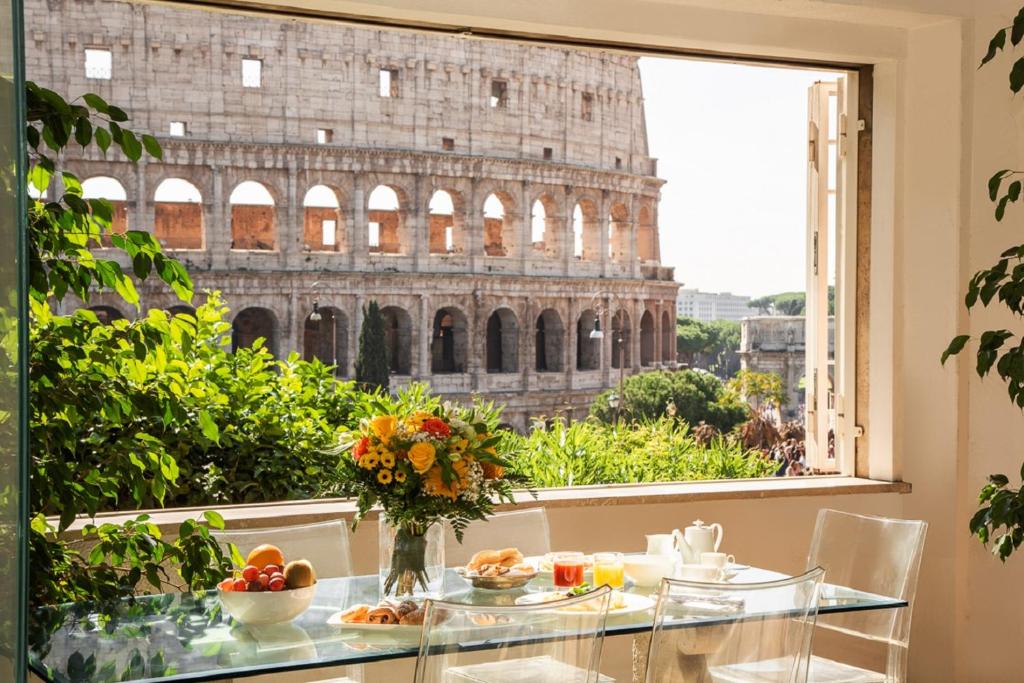 vista da sala do 47Luxury Suites - Colosseo, com café posto à mesa de vidro, cadeiras e vaso de flores, dando vista para o Coliseu e plantinhas ao redor