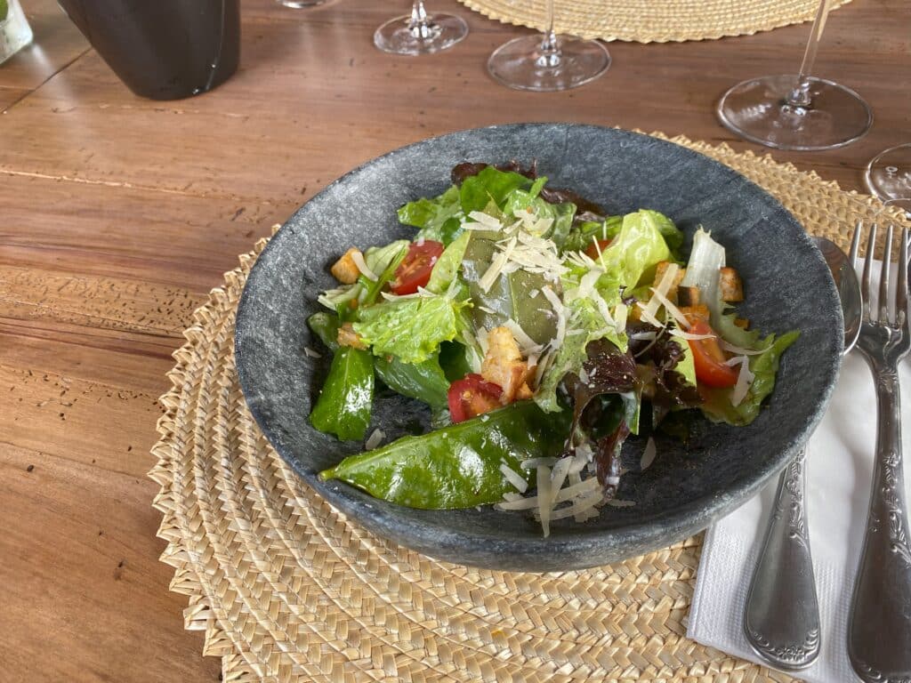 Salada Caesar do restaurante Apino, em prato fundo de cerâmica em estilo rústico, apoiado sobre lugar americano arredondado, de palha, com talheres ao lado direito