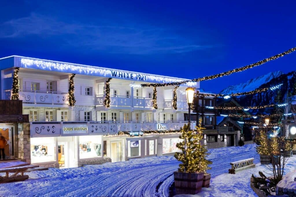 Vista de fora do hotel White 1921 Courchevel. Ele é todo branco e está iluminado com luzes de natal. No térreo é possível ver fachadas das marcas Louis Vuitton e Dior. Em frente ao hotel há uma estrada de neve e pequenas árvores também iluminadas.