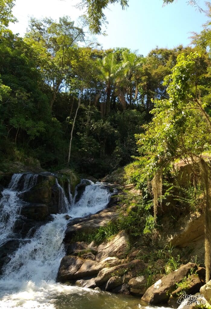 Cachoeira do Poção em Miguel Pereira, no Rio de Janeiro.  No lado direito árvores e muitas pedras, no lado esquerdo a cachoeira, em cima muitas árvores e um céu clarinho azul.