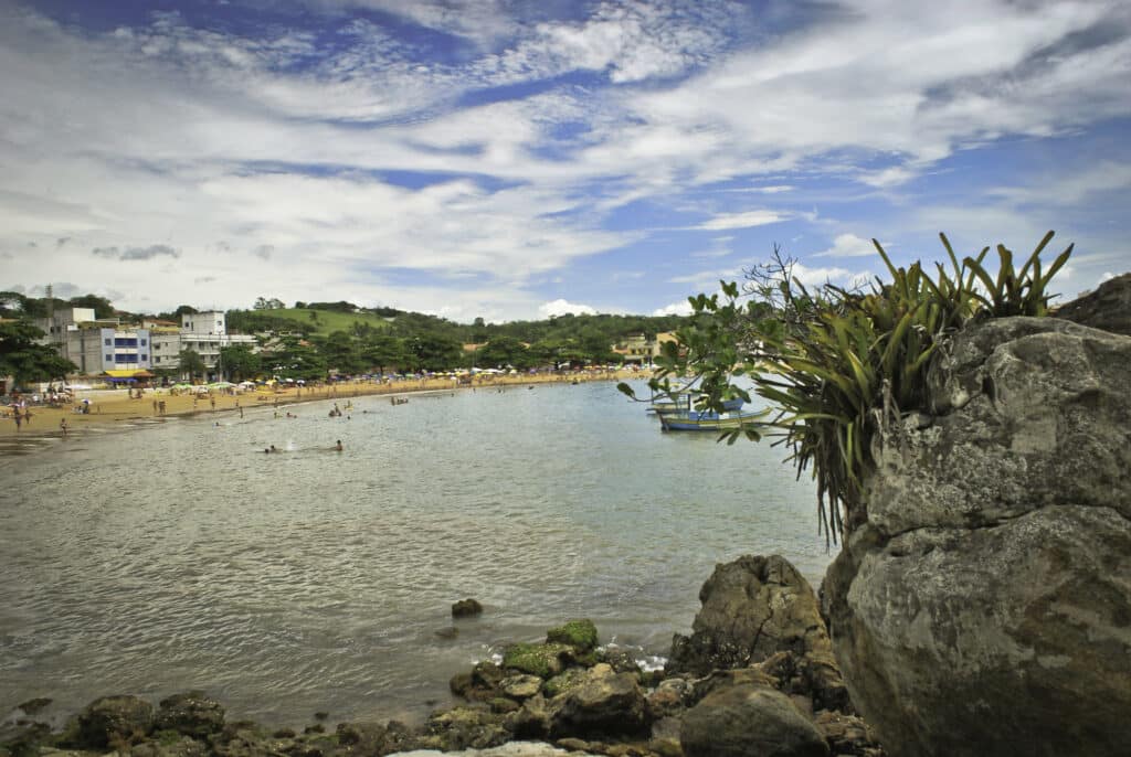 Praia de Iriri, no Espírito Santo. Uma pedra no canto direito com plantas em cima, o mar atrás e no fundo a areia com pessoas no mar.