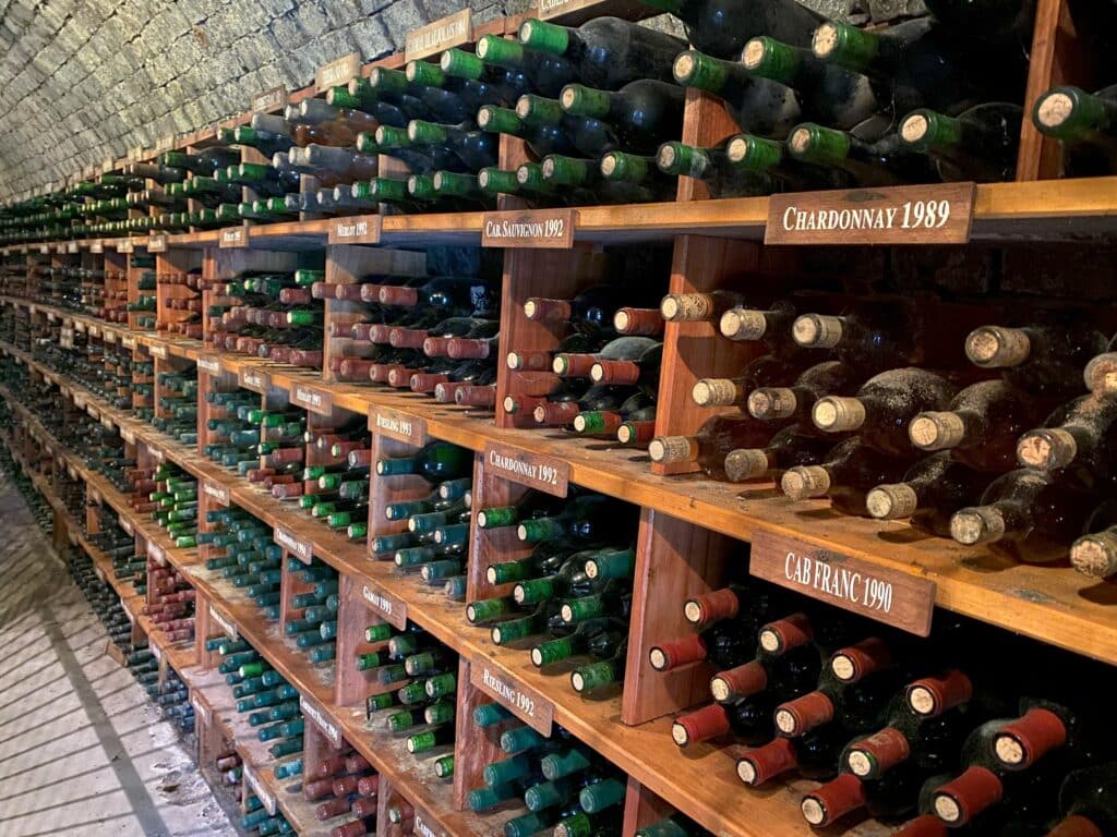 Adega com vinhos antigos diversos da vinícola Dal Pizzol. Há garrafas empilhadas em estantes de madeira com divisões, e placas indicativas mostram qual a variedade de uva dos vinhos em cada espaço. Em primeiro plano se vê um Chardonnay 1989 e, logo abaixo dele, um Cabernet Franc de 1990.