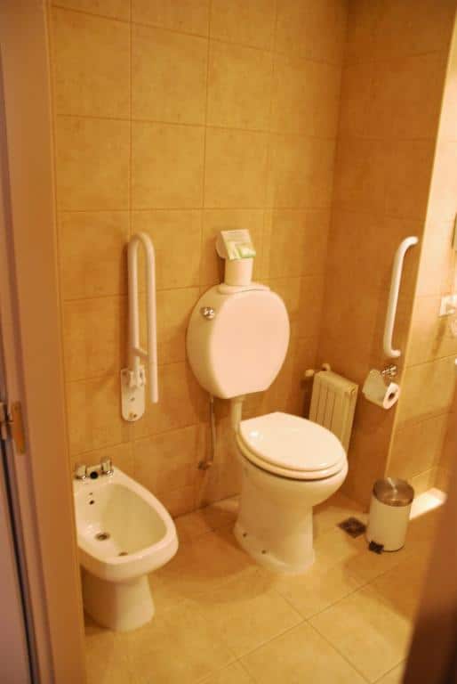 Banheiro do Alto Calafate Hotel. Um bidê do lado esquerdo, uma barra de apoio, o vaso sanitário e outra barra de apoio. Em baixo da barra, o papel higiênico e o lixo.