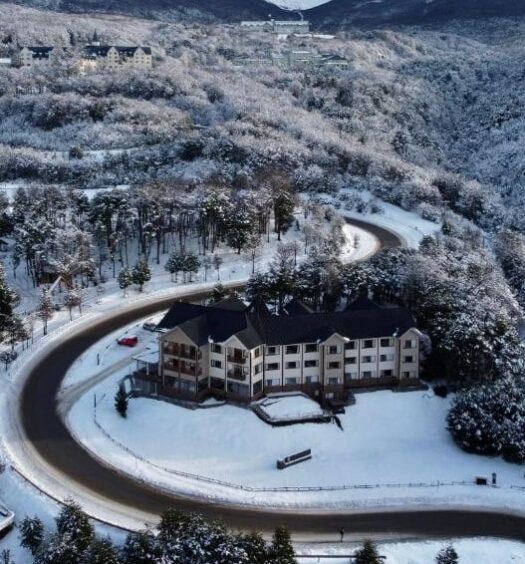 Vista de cima do Altos Ushuaia Hotel & Resto. Uma rua ao redor da propriedade, ao redor do hotel muitas árvores e no fundo as montanhas com neve. Foto para ilustrar post sobre Hotéis em Ushuaia.