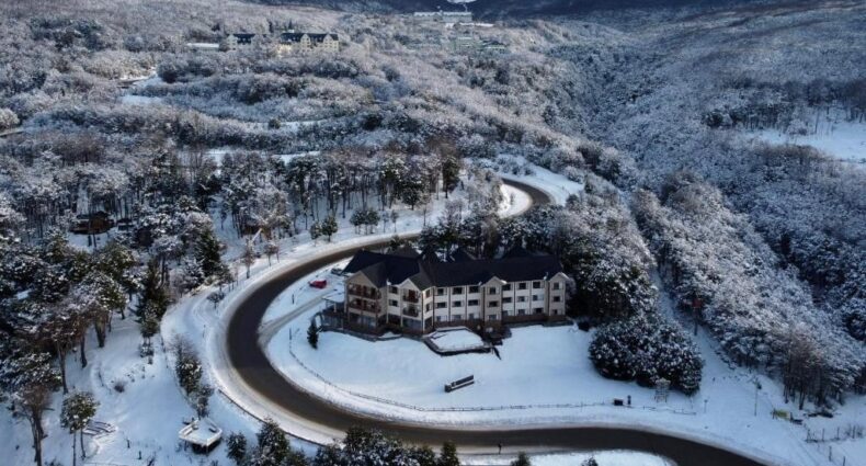 Vista de cima do Altos Ushuaia Hotel & Resto. Uma rua ao redor da propriedade, ao redor do hotel muitas árvores e no fundo as montanhas com neve. Foto para ilustrar post sobre Hotéis em Ushuaia.
