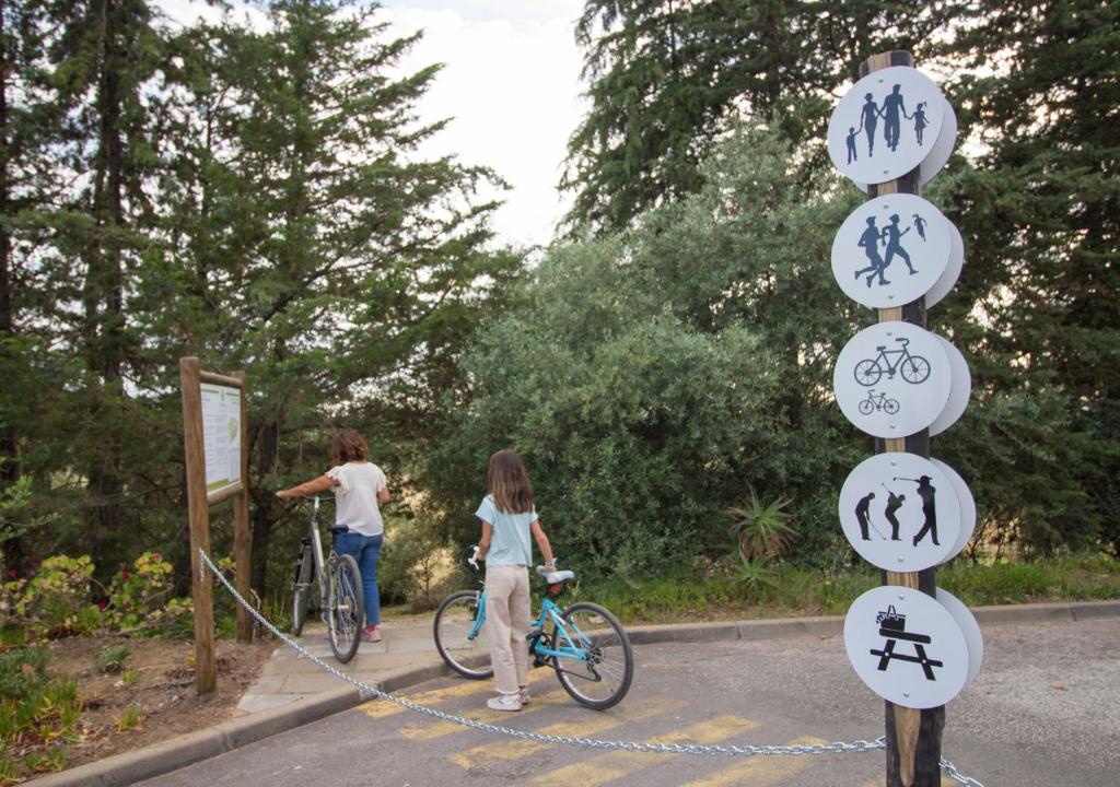Área externa do Evora Hotel. Na imagem há uma floresta ao fundo, duas pessoas ao lado de duas bicicletas e um poste com placas indicando os esportes que podem ser praticados no local.
