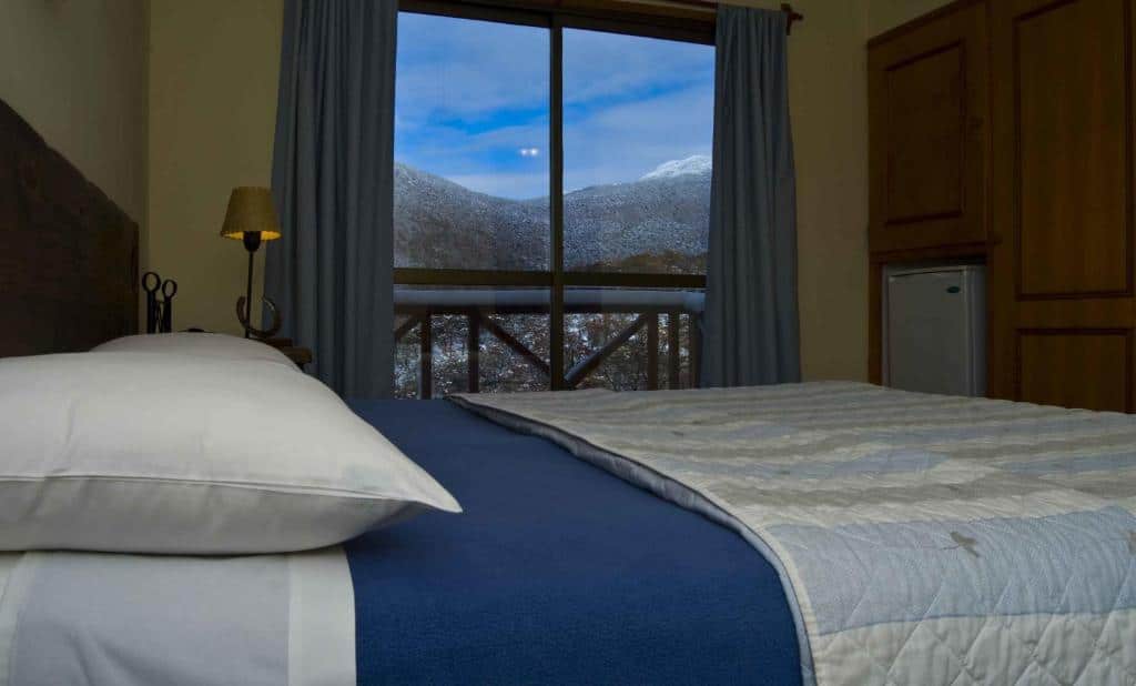 Quarto do Bagu Ushuaia Hotel. Uma cama de casal na frente, do lado esquerdo, atrás, um abajur, e do lado direito, um frigobar e um guarda-roupa. No fundo do quarto uma janela com sacada. Foto para ilustrar post sobre hotéis em Ushuaia.