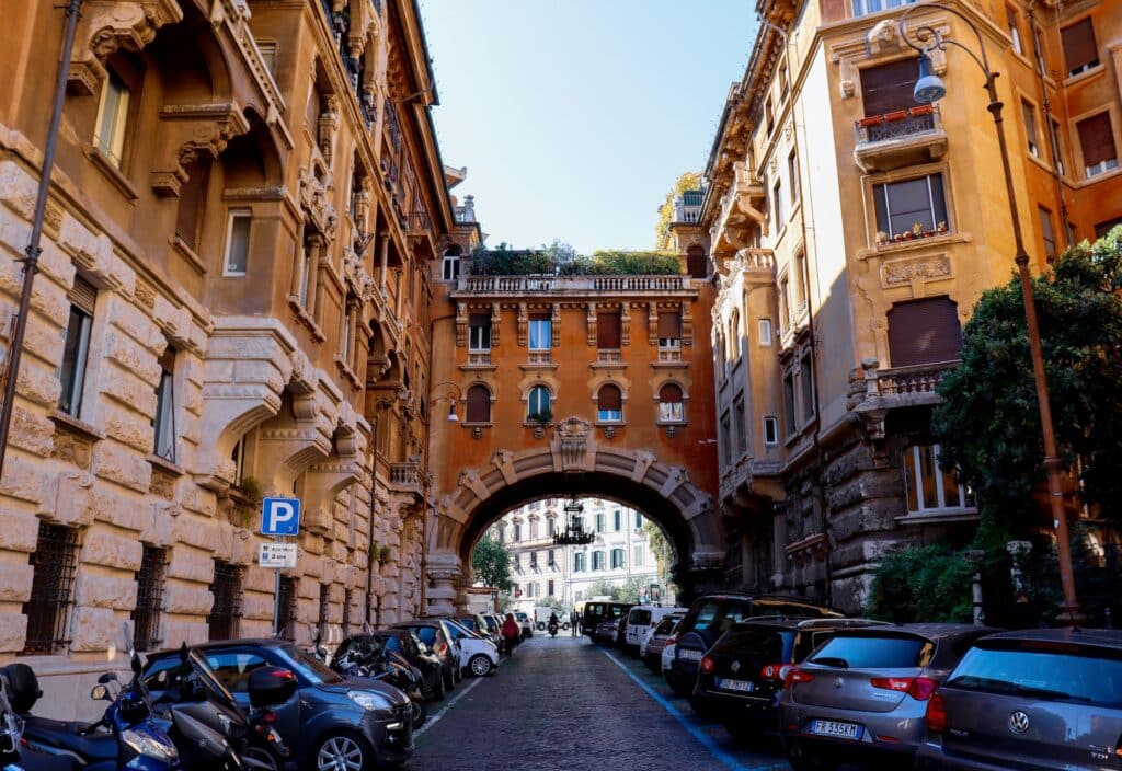 diversos carros estacionados lado a lado no Bairro Coppedé, em Roma, com um belo arco e prédios históricos ricamente trabalhados