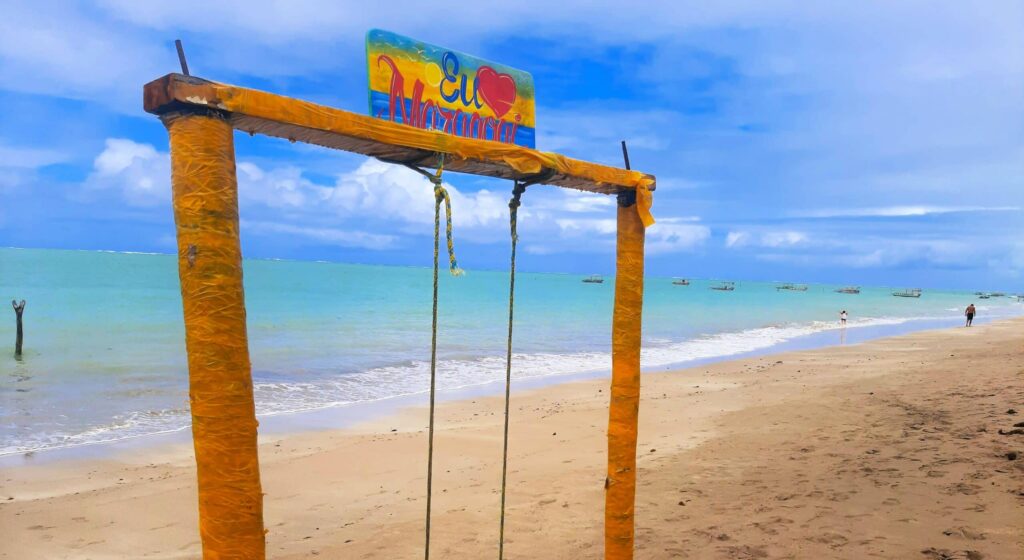 Balanço amarelho com uma placa acima escrito "eu amo maragogi", com o mar azulado ao fundo