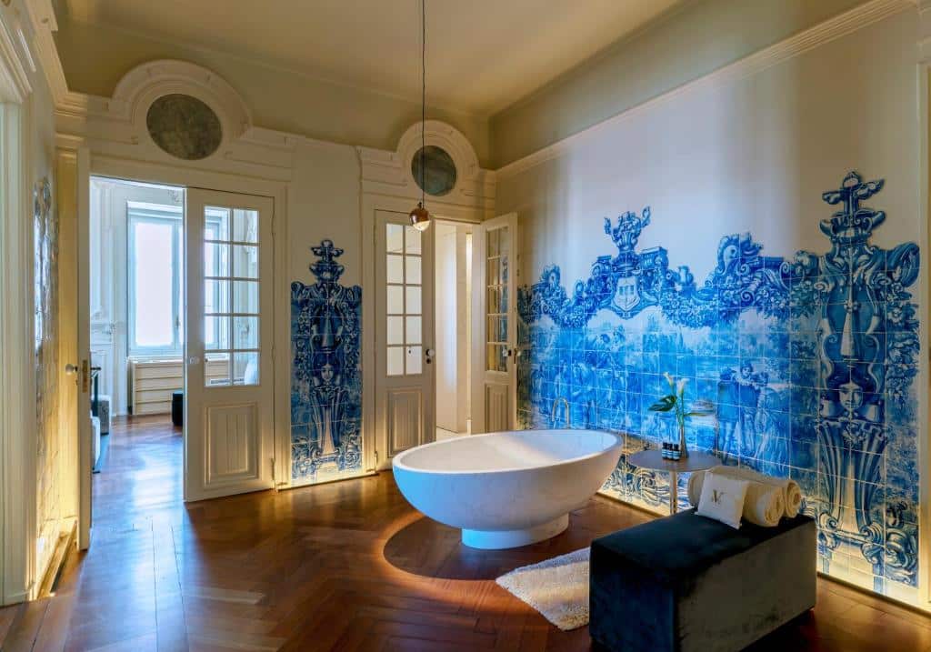 Banheiro do tamanho de um cômodo no Verride Palácio Santa Catarina, as paredes do ambiente são de azulejos portugueses azuis, no centro do local tem uma banheira oval, junto de um pequeno sofá com toalhas, para representar hotéis boutique em Lisboa