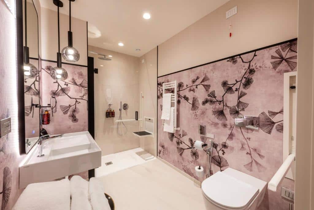 banheiro do H10 Palazzo Galla, uma das opções de onde ficar em Roma e dos melhores hotéis em Roma, com acessibilidade para pessoas com deficiência, com barras de apoio, cadeira de banho embutida, vaso mais alto e pias mais baixas em um ambiente amplo, as paredes são decoradas