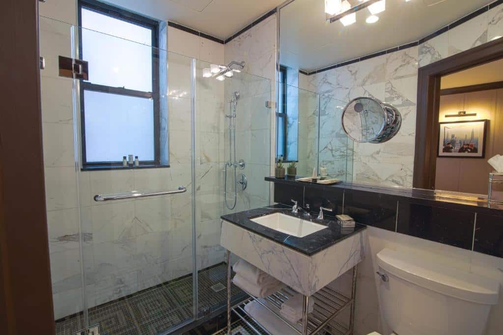 banheiro do Iroquois New York Times Square com paredes de mármore, amplo espelho no lado direito da imagem, com um pequeno espelho de aumento redondo, além de um box amplo de vidro transparente do lado esquerdo do ambiente.