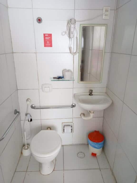 Um banheiro na Pousada Ciranda, em Itamaracá. Na esquerda vemos um vaso sanitário com ducha e barras de apoio em sua volta. Na direita há uma pia pequena, um espelho e um cesto de lixo encostado no canto.