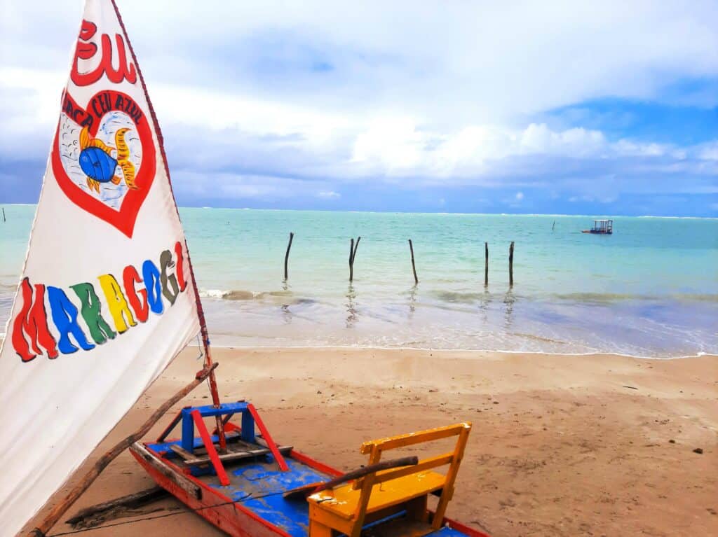 Barco na areia com uma bandeira escrito "Maragogi" com o mar ao fundo com poucas ondas, alguns galhos fincados na água e um barco navegando ao fundo