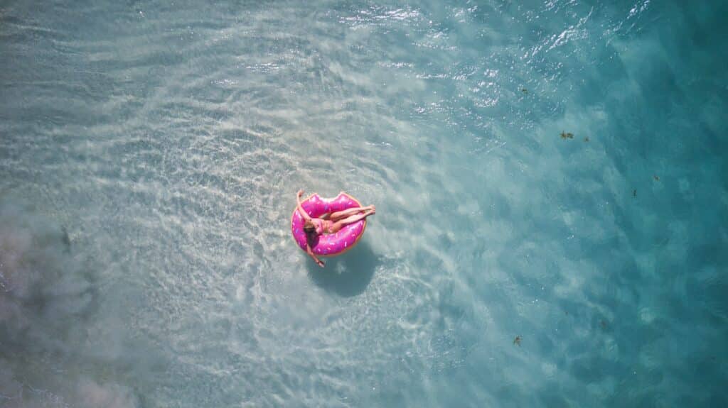 Visão de cima de uma mulher flutuando em uma boia em formato de donut cor-de-rosa no meio do mar cristalino. - Foto: Danny de Groot via Unsplash