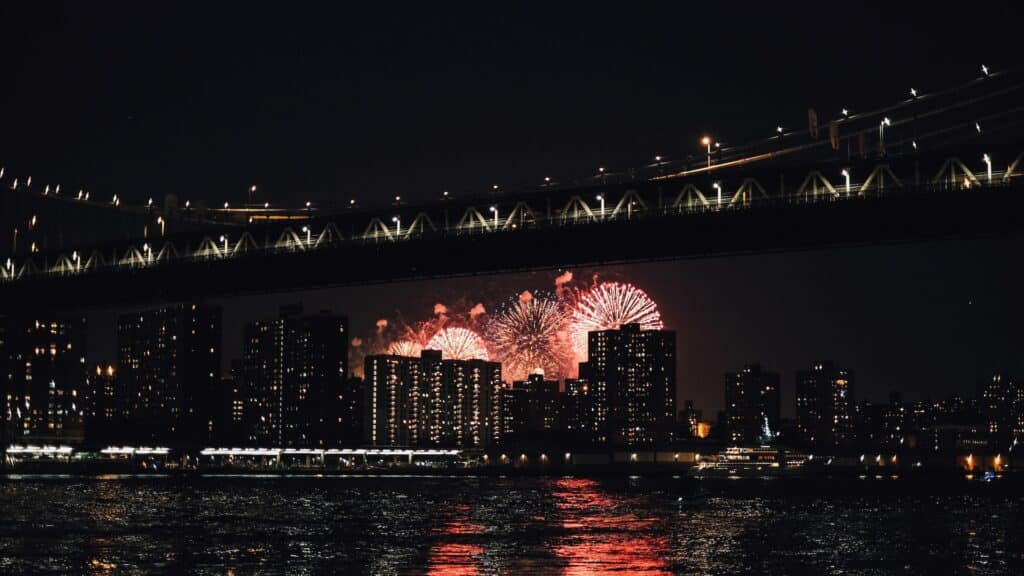 Queima de fogos em uma cidade de noite com os prédios iluminados pelos fogos na cor vermelha, de frente para a cidade há a ponte Brooklyn Bridge e o rio Hudson