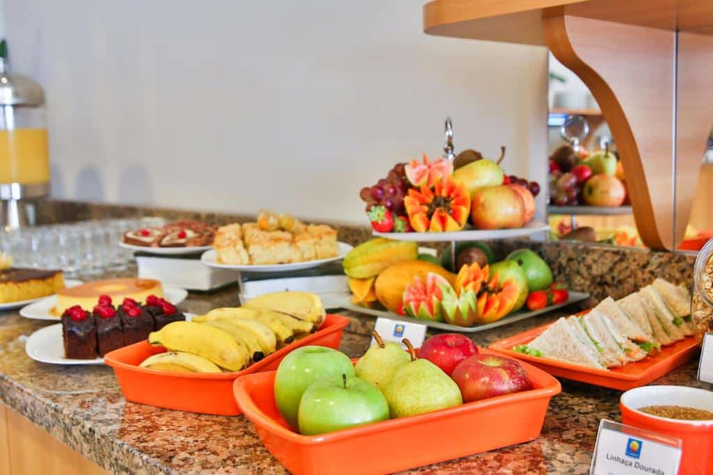 balcão de café da manhã do Comfort Hotel Joinville com várias frutas, como maçãs verdes e vermelhas, peras, mamão, uvas e demais frutas, além de pães e bolos cortados e dispostos ao redor.
