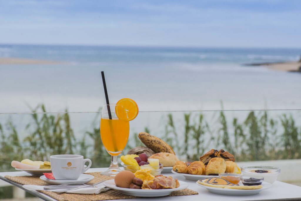 Mesa com o café da manhã do Hotel HS Milfontes Beach. Na mesa há uma xícara, uma taça com suco, pratos com pães, salgados e doces. Ao fundo da mesa é possível ver o mar.
