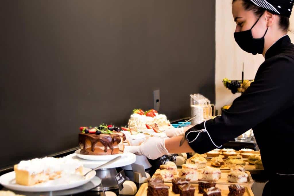 mesa de café da manhã do Naalt Hotel Joinville mostrando vários bolos confeitados, pedaços de cucas cortados sobre os pratos e uma mulher branca, usando máscara de tecido, e luvas de plástico, organizando os pratos