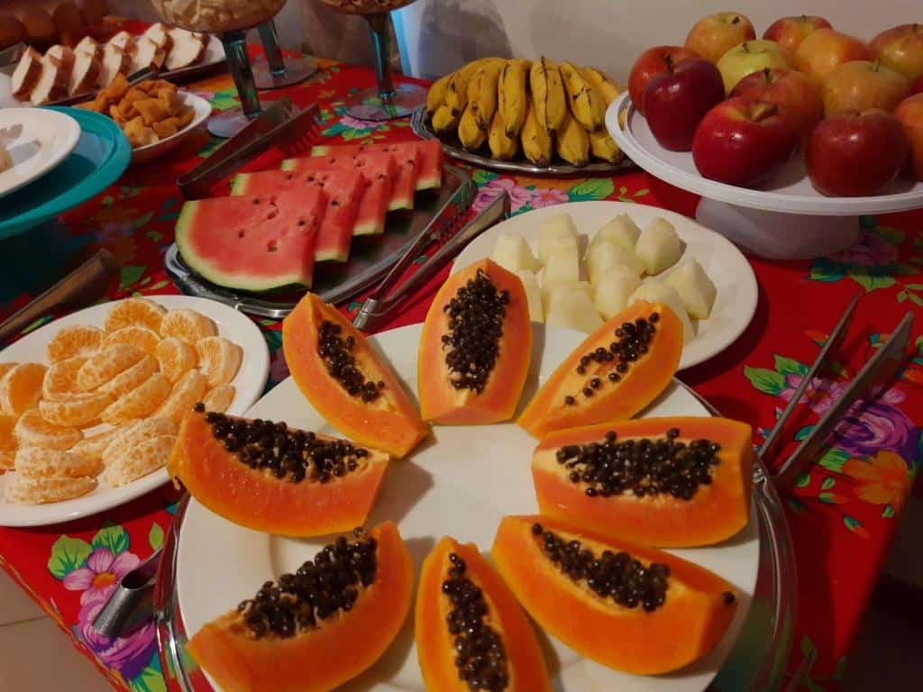 Mesa de café da manhã da Pousada Il Conventino. Podemos ver frutas como mamão. laranjas, melancias, maçãs e bananas. Outros alimentos como pães também estão visíveis no canto da imagem.