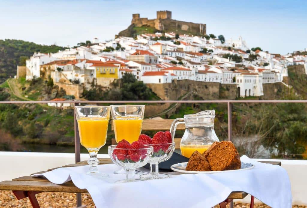 Mesa com café da manhã no hotel Quinta do Vau. Na mesa há uma jarra e duas taças de suco de laranja, duas fatias de bolo e duas taças pequenas com morango. Ao fundo da mesa é possível ver uma pequena cidade com um castelo medieval.