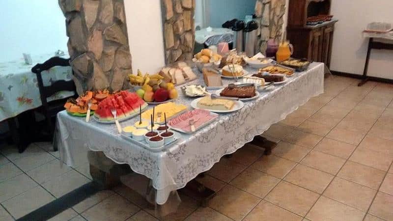 mesa de café da manhã da Schulz Pousada com vários pães, bolos e frutas picadas, além de sucos, iogurtes e café.