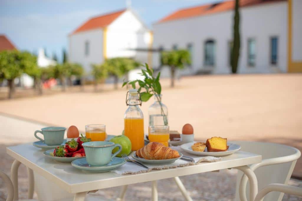 Mesa com café da manhã no Torre de Palma Wine Hotel. Na mesa há uma jarra de suco, copos, xícaras, frutas, bolo, ovos e um croissant. Ao fundo é possível ver árvores e o hotel.