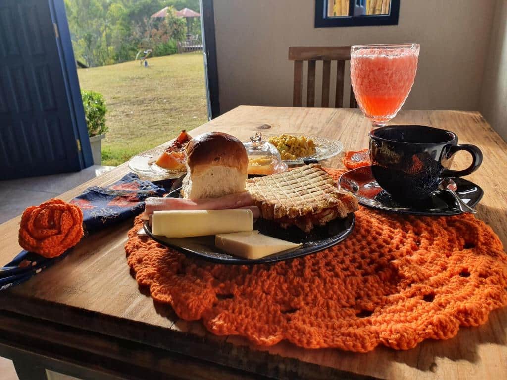 Imagem de uma mesa de café de manhã no Sítio Juara. Sobre ela vemos alimentos como pães, frios, frutas, etc. Também há xícaras e um copo com suco. No canto direito vemos uma porta que está aberta e dá passagem para a área exterior da pousada.
