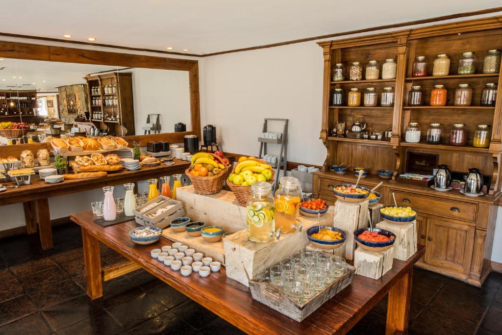 Café da manhã do Calafate Parque Hotel. Uma mesa no centro com frutas, copos e sucos. No lado esquerdo uma mesa com pães e café, em cima um espelho. Atrás, no lado direito, uma cômoda com potes de temperos e decoração.