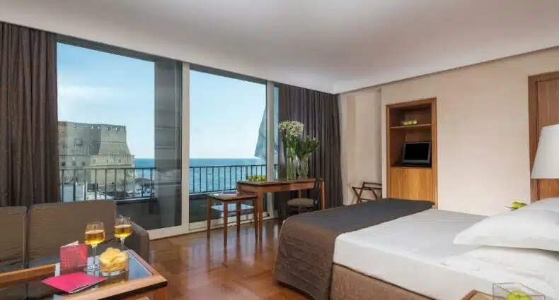 Quarto do Hotel Royal Continental com uma cama de casal, uma mesa de vidro com duas taças em cima e ao lado duas cadeiras, ao fundo outra mesa de madeira com cadeira e atrás dela uma varanda com vista para a Baía de Nápoles e para o Castelo dell'Ovo, para representar hotéis em Nápoles.