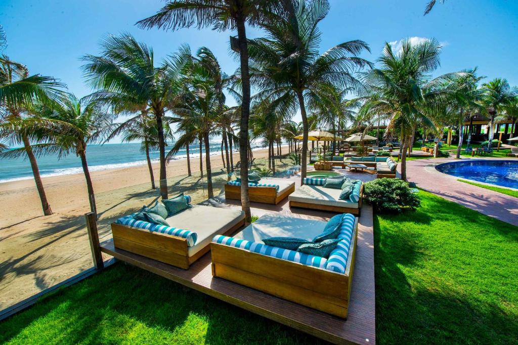 Parte do Carmel Cumbuco Resort que mostra vários sofás grandes em frente a praia com algumas palmeiras em volta e ao fundo uma piscina azul durante o dia, ilustrando post Pousadas em Cumbuco.