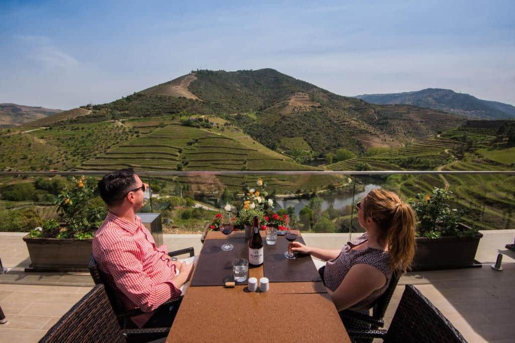 Casal sentado em uma mesa tomando vinho no Vila Gale Douro Vineyards durante o dia com vista para o rio e as vinhas.