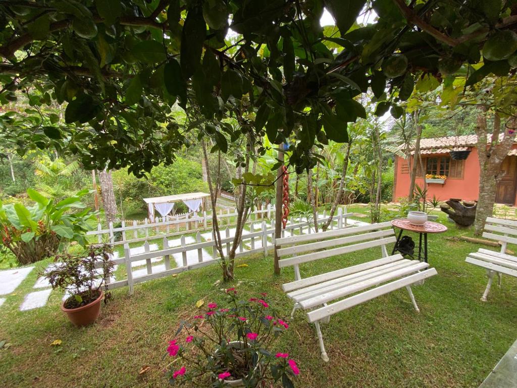 Imagem de um jardim no Chalé da Estância. Há diversos vasos com plantas e bancos sobre a grama. No fundo da foto é possível ver um deque com cortinas e uma rede de balanço.