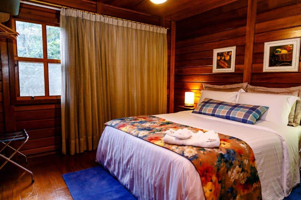 Quarto da pousada Chez Domaine Pousada Organica. A cama de casal está no meio, ao lado de uma mesinha de cabeceira. Do lado esquerdo está a janela com cortinas. As paredes são de madeira. Esta é uma das pousadas em Pedra Azul.