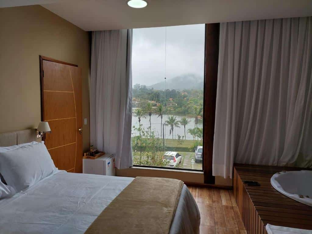 Quarto do Costa del Mar Hotel Boutique. Uma banheira de hidromassagem do lado direito, do lado esquerdo uma cama de casal, a porta do quarto e um frigobar. No fundo, uma parede de vidro com cortinas e vista para o lago.