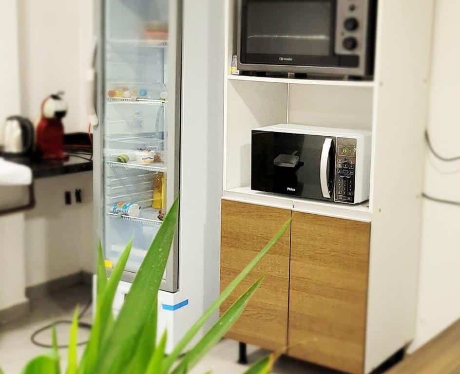 Cozinha da Pousada São José com um microondas e um forno elétrico dispostos em cima um do outro num armário de MDF. Ao lado há uma geladeira com porta de vidro transparente e alguns alimentos nas prateleiras.