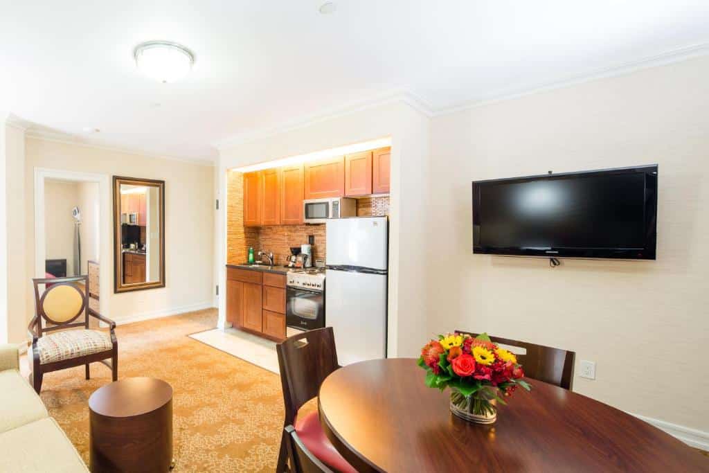 sala de estar e cozinha integrada do Radio City Apartments com uma de jantar redonda e de madeira no lado direito da imagem, abaixo de uma televisão de parede. No lado esquerdo é possível ver uma pequena cozinha com armários de madeira, pia, microondas, cafeteira elétrica, fogão a gás e uma geladeira.