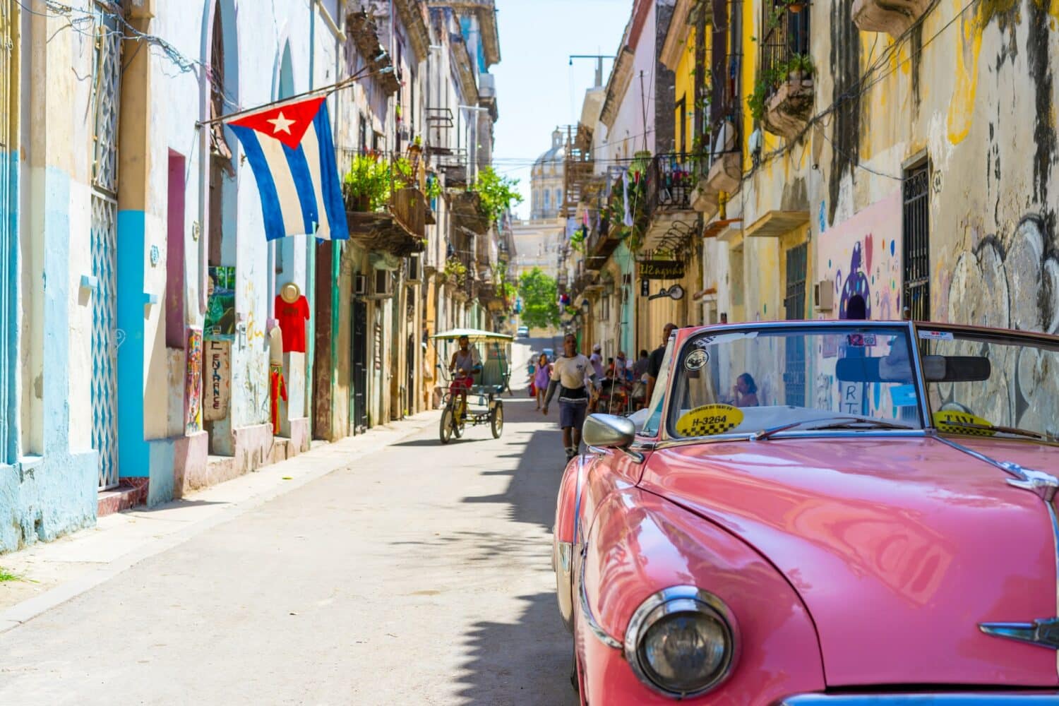 Rua em Havana para ilustra o post sobre seguro viagem Cuba. No canto direito fica um conversível vermelho, e ao fundo várias casinhas coloridas ladeiam a rua asfaltada. Algumas pessoas caminham por ali, e um homem anda de charrete no meio do local. No canto esquerdo mais ao fundo está uma bandeira de Cuba. - Foto: Alexander Kunze via Unsplash