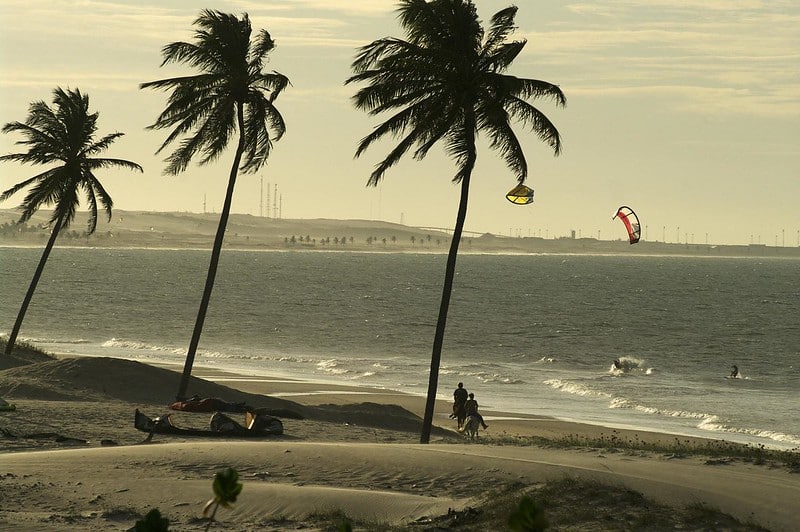 Praia de Cumbuco com três palmeiras, duas pessoas andando em cavalo, duas pessoas praticando kite surf na água e muita areia em volta durante o dia, ilustrando post Pousadas em Cumbuco.