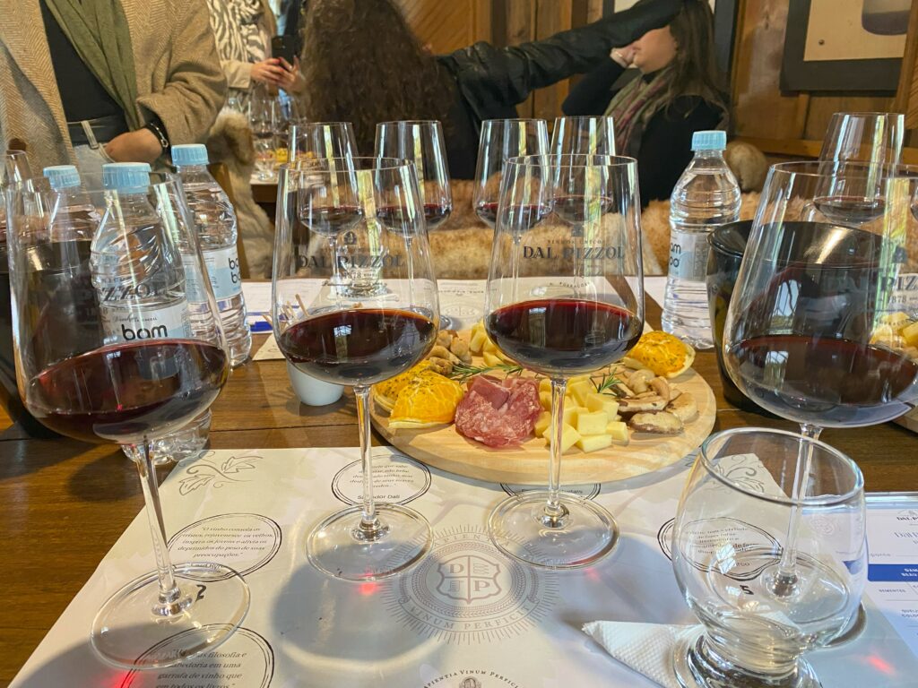 Quatro taças altas com vinhos tintos da Dal Pizzol em degustação harmonizada da vinícola, com tábua de frios e queijos ao fundo, ao lado de garrafas de água