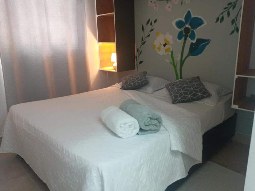 Cama de casal num quarto dos Dominican Dreams Apartments, um dos hotéis perto do aeroporto de Punta Cana. Há duas toalhas enroladas aos pés da cama, e apoios de cabeceira dos dois lados, sendo que o do lado esquerdo tem um abajur aceso. Há uma pintura de flores na parede atrás da cama.