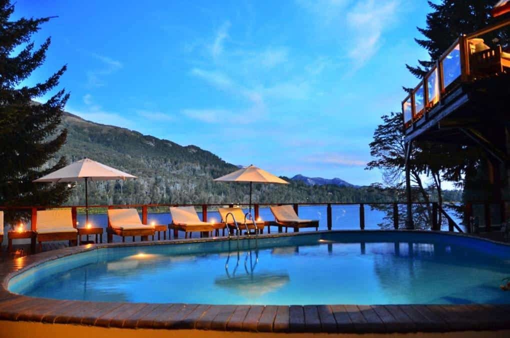 Piscina ao ar livre do Dos Bahias Lake Resort. Uma piscina na frente, atrás guarda-sóis e cadeiras de tomar sol, com um vidro cercando e vista o lago e as montanhas.
