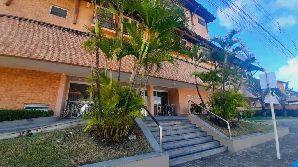 Fachada do hotel Ponta Verde Praia do Francês, com uma escada e jardim dos lados, e ao fundo o prédio com bicicletas na frente e carrinhos para carregar malas. Há quartos com sacadas